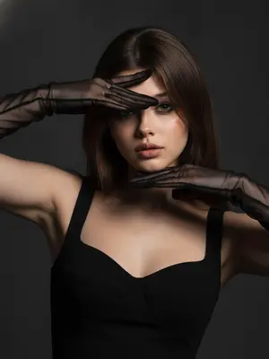 На фотографии тесным кадром запечатлена очаровательная женщина на темно-сером фоне, одетая в прозрачные черные перчатки и черное платье. Она элегантно позирует, обрамляя свое лицо руками, создавая игривые тени вокруг глаз. Ее яркий глазной макияж с аккуратной подводкой, безупречная кожа, кинематографическое освещение и высокомодный стиль добавляют атмосферу загадочности и соблазнительности.