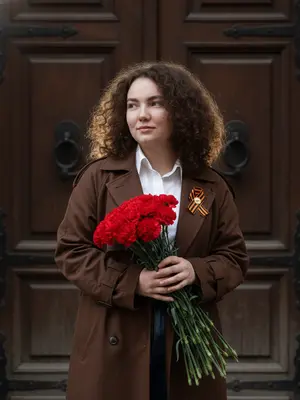 На улице, перед массивными дверями из старинного дерева, запечатлена модель в элегантной позе. В руках у нее букет красных гвоздик, а взгляд устремлен в сторону с легкой полуулыбкой на лице. Ее тело слегка повернуто, создавая уверенную атмосферу. Фон четкий, не размытый, а свет играет по контурам ее силуэта, придавая изображению живость.

Портрет снят на iPhone 16 Pro Max в стиле стритстайл с эстетикой тихой роскоши. Камера расположена на уровне груди, создавая натуральную перспективу. Кожа модели изображена максимально реалистично - видны поры, текстура, микродетали, и легкая неоднородность тона придают ей естественность. На лице нет искусственного сияния, а лишь мягкое влажное отблеск, который придает коже живой вид.

Фотография обработана минимально, сохраняя натуральные цвета и высокую детализацию текстур. Никаких фильтров или искусственных артефактов - лишь реалистичное отражение модели и окружения.