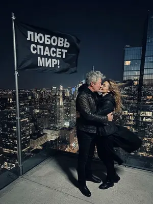 Изображение: Молодая пара, одетая в черное, стоит на вершине высокой башни, обнявшись и целуясь. Они стоят против ветра, который развевает их волосы и одежду. Рядом с ними развевается черный флаг с надписью «ЛЮБОВЬ СПАСЕТ МИР!». Вокруг них виднеется городской пейзаж с высокими зданиями и огнями, отражающимися в стеклянных окнах. Мужчина и женщина выглядят решительными и страстными, их лица освещены лунным светом, подчеркивающим контуры их фигур и выражение лиц.