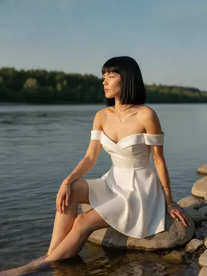 Женщина сидит на гладких камнях у реки, где вода плавно шуршит. Ее тело немного повернуто к воде, одна нога изящно согнута, ступни касаются воды. Взгляд устремлен вдаль, лицо выражает задумчивость и спокойствие. Фотография вертикальная, сделана на уровне глаз с небольшим наклоном. На шее блестит тонкая цепочка с кулоном, а на запястье носит минималистичный браслет.