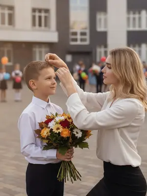На фотографии, посвященной 1 сентября, запечатлен трогательный момент у входа в школу. Сбоку виден первоклассник, который гордо держит в руках яркий осенний букет, наполненный цветами. Ребенок с доброй улыбкой смотрит на свою маму, слегка приподняв подбородок. Мама, стоя перед ним, наклонилась вперед и заботливо поправляет его прическу, используя обе руки. Ребенок стоит с расслабленной осанкой, держа букет перед собой, создавая атмосферу нежности и радости в этот особенный день.