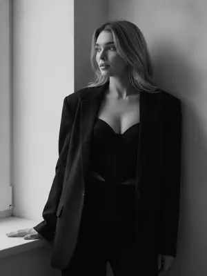 Женщина стоит в углу комнаты, наклонившись к стене. Ее лицо, с выражением спокойной уверенности, обращено к окну, вдалеке. На ней большой черный пиджак, небрежно надетый на плечи, и топ-корсет с V-образным вырезом. Рука лежит на пиджаке, поддерживая ее. Комната пронизана естественным светом из больших окон слева. Общая атмосфера внутри - стильная и спокойная, подчеркивающая элегантность и минимализм. Женщина позиционируется справа от центра кадра. Освещение мягкое, придающее ощущение простоты и минимализма. Черно-белая палитра фотографии выделяет контрасты и текстуры, создавая загадочную, но элегантную атмосферу. Портрет выполнен в ультрареалистичном 8K разрешении.