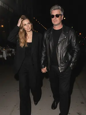 Capture a natural, paparazzi-style image under the night sky. A couple strolls together in the dimly lit street. The woman leads the way, casually adjusting her hair with a gentle gesture, while the man follows a bit behind, hand in hers. The camera captures the scene from a slightly elevated angle, zoomed in closely to the pair. Maintain the essence of a high-end celebrity magazine photograph. The facial features, eyes, nose, lips, hairline, and skin texture must stay true to their original form without any artificial alterations.