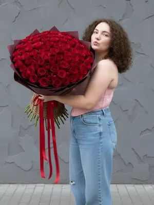 Женщина стоит наполовину повернутая, с наклоном головы и улыбкой, слегка скривив губы в форме «бантика», отправляя воздушный поцелуй. На ней корсет нежно-розового оттенка с объемным узором, напоминающим лепестки, в сочетании с нежно-голубыми свободными джинсами, прямого кроя, немного мешковатыми и украшенными потертостями. Ее кожа лучится, обладая легким загаром. В ее руках букет цветов, где присутствует удивительный эффект отражения, создающий впечатление двойственности.