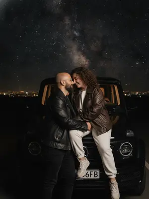 Ночной снимок пары на фоне черного Mercedes-Benz. Снимок сделан сбоку, где женщина удобно расположилась на капоте, а мужчина стоит рядом, нежно касаясь ее ноги. Их лица близки друг к другу, носы почти соприкасаются, глаза закрыты, и на губах играет легкая улыбка. Она обнимает его за шею, создавая атмосферу интимности и романтики. Ноги девушки переплетены за спиной партнера, что добавляет динамики в композицию. Камера фиксирует момент на уровне груди, в среднем плане. Снимок сделан на Sony A7 IV в формате RAW, с использованием объектива 50mm, ISO 800 и диафрагмы f/1.8. Яркая вспышка освещает сцену, создавая мягкий теплый свет от автомобиля и глубокие тени вокруг.