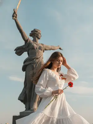Фотосъемка возле монумента «Родина-мать зовет» в Волгограде. Камера смотрит вверх, немного наклонена. Женщина в кадре, вращается вправо. Волосы, ткань и пальцы с небольшим размытием от движения. Голова опущена, глаза закрыты, выражение спокойное. Рука у головы, расправляет прядь. Другая рука держит красный цветок. Белое платье с длинными рукавами, V-образным вырезом, рюшами и воздушной юбкой, развевается. На груди - Георгиевская лента. Вдалеке монумент «Родина-мать зовет» - женщина с мечом и поднятой рукой, голова повернута. Небо голубое, с облаками. Солнечный свет, теплые отблески, мягкие тени. Женщина справа внизу, монумент вверху. Цветокоррекция естественная, высокая детализация.