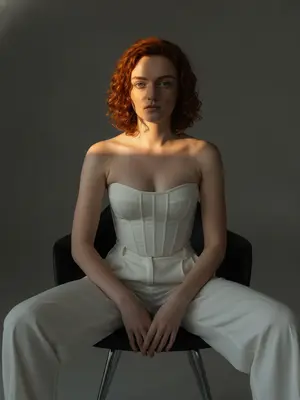 Женщина, излучающая стиль и уверенность, запечатлена на фотографии в жанре моды. Она занимает расслабленную позу, усевшись на современном черном кресле. Ноги разведены в стороны, торс слегка наклонен вперед, а предплечья лежат близко к коленям. Руки свободно соединены перед собой, создавая гармоничный образ. Фон студии выполнен в минималистичном темно-сером стиле, лишенном лишних деталей, что придает снимку сдержанность и элегантность.
