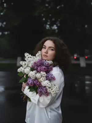 На темном фоне ночного города, тенистые деревья и майская листва создают задний план. Женщина стоит перед камерой, обе руки обнимают огромный букет сирени, белой и фиолетовой. Ее голова слегка наклонена к цветам, словно вдыхая их аромат. Макияж подчеркивает глаза черными стрелками и длинными ресницами, на губах блеск. Съемка происходит сбоку на уровне глаз, при слабом ночном освещении. Изображение высокой детализации, снятое на iPhone 17 Pro, передает живой момент с легким цифровым шумом и зернистостью, без использования вспышки.