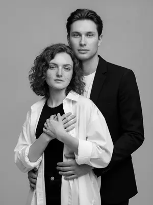 Черно-белый портрет мужчины и женщины в стиле модной фотосессии в студии. Сохраняйте их выражения лиц. Фон мягкий и однородный, светло-серый, создающий интимную и спокойную атмосферу. На переднем плане находится женщина, окруженная приглушенными оттенками серого, подчеркнутыми студийным светом, который ласкает их кожу и одежду. Ее руки лежат на груди мужчины, который нежно обнимает ее сзади, положив свою руку поверх ее.