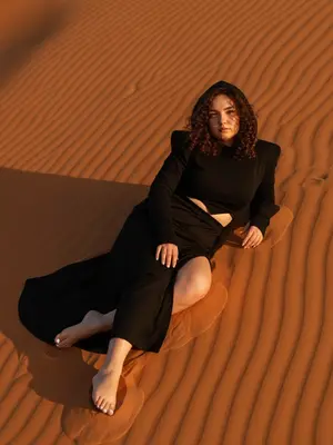 In the soft glow of the golden hour, a woman reclines gracefully on the undulating surface of a sun-kissed orange sand dune. The full view captures her from head to toe, with both feet elegantly displayed, the scene captured from a low angle. Her body is positioned diagonally across the dune, one arm supporting her as the other hand casually rests by her thigh. With one leg extended and the other slightly bent, the high slit of her skirt adds a touch of allure to the ensemble.

Gazing directly at the camera, her expression exudes a sense of calm confidence. She is adorned in a striking black hooded ensemble, featuring structured shoulders, form-fitting long sleeves with thumbholes, a waist cutout, and a floor-length skirt with a daring slit. The makeup is kept minimal, creating a dewy look that enhances her natural beauty. Her clean white manicure and realistic skin texture, complete with visible pores, add to the authenticity of the image.

The warm, golden light bathes the scene, casting gentle shadows and highlighting the intricate textures of the sand and fabric. Shot with a Canon EOS R and a 24-70mm lens, the focus is sharp, capturing every detail with a hyper-realistic quality. The overall atmosphere is cinematic yet natural, evoking a sense of editorial elegance in the desert setting.