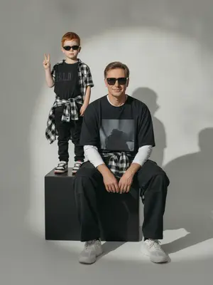 A photorealistic studio portrait captures a minimalistic editorial vibe, showcasing two individuals in a static scene. The model is seated on a large matte black cube, body slightly turned towards the camera, with knees bent and feet resting on the floor. Their arms are relaxed in front of their knees, fingers neatly extended. Standing behind and to the left on the same cube is a child, exuding confidence and playfulness. The child raises one hand in a "V" gesture while the other hand rests by their side, both figures sporting narrow dark sunglasses and directing their gaze towards the camera.

The composition features a frontal angle without noticeable perspective distortion, with the lens capturing a normal/short portrait view. The aperture is set around f/5.6–f/8, providing a moderate depth of field that keeps both subjects sharp while slightly softening the background. Soft studio lighting from a key source on the front left creates smooth transitions and gentle shadows against the backdrop, enhancing the overall aesthetic.

The background is a seamless light cyclorama with a prominent oval light spot in the center, surrounded by a subtle dark vignette. The studio setting is minimalistic, devoid of props, emphasizing a calm and stylish ambiance with a hint of retro street fashion and familial warmth. The seated model wears a dark oversized T-shirt with a bold print, layered with a light longsleeve and wide cargo trousers. They tie a plaid shirt around their waist and don chunky light sneakers. The child's attire mirrors the dark color scheme, with a T-shirt featuring a prominent chest inscription, paired with dark trousers, a plaid shirt, and colorful sneakers.

Details such as realistic fabric textures, natural creases, and fine folds add depth to the clothing, while the skin texture appears natural and unaltered. The scene is captured with high detail and sharpness, reminiscent of film photography with a light grain effect. The overall look is characterized by moderate contrast, a soft matte finish, and a focus on authenticity, free from overexposure or distracting elements.