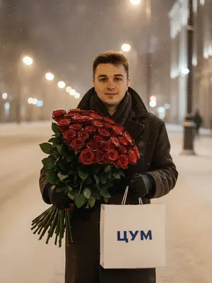 Мужчина в темно-коричневом пальто, шагает по улице, покрытой снегом, под ночным небом. В его руках громадный букет красных роз с зелеными листьями и пакет отмеченный логотипом "ЦУМ". Его взгляд сосредоточен, излучая уверенность и спокойствие, с легкой улыбкой на лице. Вспышка освещает его лицо и аксессуары, создавая игру света и тени, подчеркивая контуры лица и верхней одежды. На заднем плане пространство города, покрытое снегом, с приглушенными огнями уличных фонарей и пустыми улицами, что придает ощущение уединения и стиля. Съемка выполнена на объективе 85 мм с диафрагмой f/1.4, с акцентом на лице и аксессуарах мужчины.