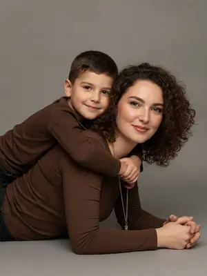 Create a studio portrait capturing the essence of a mother and child, maintaining their original facial features and expressions with precision. The scene is set in a close-up perspective at eye level. The duo is depicted lying on their stomachs, facing sideways to the camera. The child lovingly embraces the mother from behind, their arms wrapped tenderly around her shoulders. Both subjects gaze directly at the viewer, establishing a heartfelt connection. 

The mother exudes a natural beauty with a flawless, matte complexion complemented by subtle makeup - soft pink lips and a hint of mascara. She is dressed in a snug brown long-sleeved top, accentuated by a graceful silver chain and pendant draping elegantly along her collarbone. 

The child mirrors the mother's attire with their own brown long-sleeved top. They wear a delicate, thin chain necklace that harmonizes with the mother's jewelry, adding a touch of visual unity to the composition. 

The image is formatted in 9:16 ratio and rendered in stunning 8K quality.