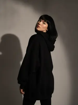 На съемке haute couture запечатлена стильная женщина, стоящая спиной к наблюдателю. Ее голова наклонена назад, взгляд устремлен прямо в объектив. Легкая улыбка играет на ее губах. Фотография сделана на 35-мм пленку с высоким контрастом, создавая жесткое драматическое освещение. Резкая тень отчетливо отражается на простом сером фоне, придающем образу эстетику 90-х. Теплая коричневая цветовая гамма добавляет кинематографичности и стиля, характерного для фэшн-журналов.

Изображение характеризуется максимальным фотореализмом, чистой редакционной фотографией с высоким контрастом и четкими контурами силуэта. Естественная анатомия, реалистичные руки без лишних пальцев, никаких дополнительных конечностей или дублированных частей тела. Лицо не искажено, не имеет деформаций. Нет запутанных нитей, сеток, текста или водяных знаков. Фотография выполнена в цвете.