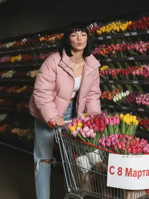 На фотографии изображена женщина, толкающая тележку, переполненную яркими цветами. На тележке висит табличка "С 8 Марта". Она одета в большой пушистый пуховик нежно-розового цвета, расстегнутый, голубые джинсы с потертостями и топ. Ее макияж безупречен, подчеркивающий естественную красоту. Женщина наклоняется вперед над ручкой тележки, с улыбкой, словно посылая воздушный поцелуй в сторону камеры. Вокруг нее полки, уставленные теми же яркими цветами, создают живописный фон. 

Фотография захватывает непринужденный и спонтанный момент. Освещение жесткое, с яркой прямой вспышкой, создающей сильные тени, пересвеченные участки и неравномерную экспозицию. Композиция наклонная, объекты не по центру, конечности обрезаны, что добавляет динамики сцене. Фокусировка неидеальна, сцена запечатлена в движении, застывшем в середине эмоций. На фотографии присутствует шум и зернистость, не скрытые ретушью, придающие ей более небрежный и документальный вид. Атмосфера на снимке интимная и реалистичная, словно мгновение из жизни, запечатленное без украшательств.