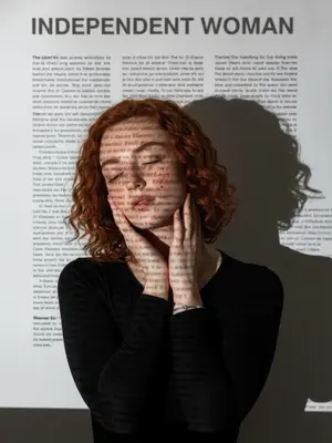 На фотографии запечатлен человек в обтягивающем черном лонгсливе с длинными рукавами и серебряным браслетом на левом запястье. Полупрофиль взгляда, голова слегка наклонена, глаза закрыты. Правая рука лежит на правой щеке, а левая опускается к основанию шеи, пальцы легко касаются линии челюсти. Фон заполнен контрастной проекцией: черная типографика "INDEPENDENT WOMAN" и колонки текста отбрасываются на белую плоскость и кожу. Образуется сложное переплетение теней и четких букв. Атмосфера сдержанная, с акцентом на гиперреалистичную текстуру кожи и мельчайшие детали без изменения оттенка. Съемка выполнена на камеру Sony, используя объективы 35mm и 85mm для создания профессиональной глубины портрета.