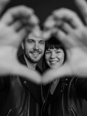 A candid and joyful couple stands out in a dreamy and intimate setting, exuding a romantic vibe that feels straight out of a movie scene. Their genuine smiles radiate warmth as they gaze directly at the viewer, creating a sense of connection. In the forefront of the image, the couple's intertwined hands subtly blur into a heart shape, delicately framing their beaming faces, adding a touch of sweetness to the overall composition.