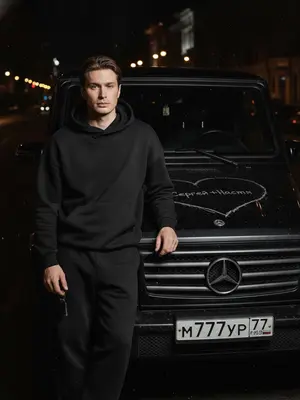 На заднем плане мрачное ночное окружение городской улицы, смутные очертания зданий и фонарей. В центре кадра человек, одетый в черный спортивный костюм без излишних деталей, стоит у автомобиля, наклонившись на капот. В руке у него легко покачиваются ключи от машины. На капоте, занимающем всю площадь кадра, выгравировано грубое сердце с таинственным текстом внутри. Отражение жесткого прямого света создает резкие тени и подчеркивает микроскопические дефекты краски, словно пыль и царапины. Съемка выполнена с верхнего ракурса, придающего кинематографическую атмосферу. Реалистичные текстуры кожи и ткани костюма подчеркиваются объективом 35 мм с диафрагмой f/2.8, создавая грубую уличную эстетику.