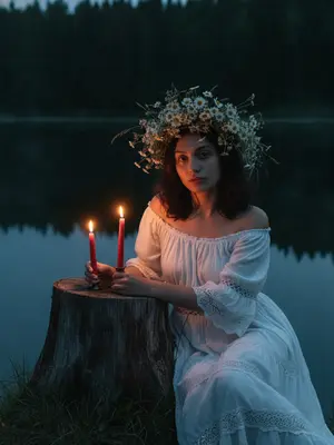 In the tranquil twilight by a serene lake, a woman is seated next to an ancient tree stump. Her posture exudes a dreamy relaxation, leaning slightly forward, one arm casually draped over her knee, the other hand delicately cradling a slender crimson candle beside another flickering flame on the stump. She wears a vintage off-shoulder peasant dress in white, adorned with intricate lace accents and gentle fabric drapes. Her hair is styled with a natural softness, featuring loose strands framing her face.

Adorning her head is a lavish crown of wildflowers, crafted from chamomile blooms, meadow grass, and delicate white blossoms. The background is enveloped by a dark forest and the placid waters of the lake, while the warm glow of the candles casts a gentle light on her face and hands, creating deep blue shadows in the mystical ambiance of a summer night.

Captured with the night mode of an iPhone, the image boasts a realistic grain in low light, with adjusted exposure to maintain sharpness and clarity, devoid of flash, blur, or bokeh effects.