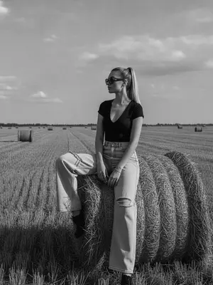 На фотографии в стиле high-fashion editorial, женщина сидит на большом тюке сена в сельской местности. Ее корпус слегка повернут, одна нога свободно опущена, а другая согнута, создавая расслабленную, но выразительную позу. Руки лежат естественно на колене и поверхности тюка. Взгляд направлен в сторону, профиль лица выражает спокойствие и уверенность. Женщина одета в черный топ с V-образным вырезом и объемные светлые джинсы с потертостями. На ней крупные солнцезащитные очки и минималистичные украшения. Фон состоит из открытого поля с несколькими тюками сена, создающими перспективу. Линия горизонта находится ниже центра кадра, подчеркивая небо. Освещение жесткое, натуральное, смягченное облаками, создающее выразительные тени и контраст. Стиль фотографии кинематографичен, с пленочным зерном и высокой детализацией. Редакционный fashion стиль акцентирует внимание на атмосфере и пространстве кадра.