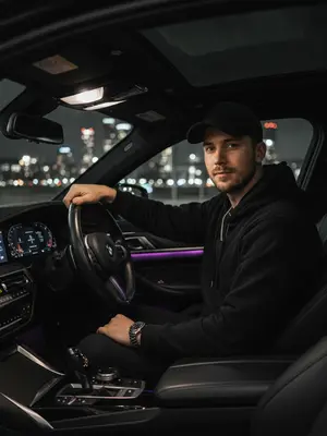 Мужчина с элегантным образом за рулем роскошного BMW, стоит на фоне ночного города. Его рубашка тщательно заправлена, подчеркивая уверенность и стиль. Серебряные часы на запястье и цепочка на шее добавляют шик. Ладонью касается руля, взгляд направлен прямо в объектив с уверенностью. Салон автомобиля пропитан атмосферой роскоши: кожаные сиденья и яркие фиолетовые светодиоды создают уют. Вдали сверкают огни городской ночи, придающие образу стиль, статус и мощь. Съемка осуществлена на объективе 50mm f/1.8 с использованием вспышки, игра света и тени придают изображению выразительность.