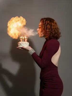 На фотографии, которая кажется почти фотокопией реальности, запечатлено удивительное событие, происходящее на празднике. Человек, изображенный в профиль, держит в руках крошечный белоснежный торт-бенто с мерцающими свечами. Из уст человека вырывается облако белой сахарной пудры, направленное к пламени свечей, вызывая внезапное вспышечное пламя, похожее на шар. В воздухе над тортом смешиваются пламя и частицы белой пудры, создавая удивительный эффект. Фон студии серый и нейтральный, с резкой тенью, добавляющей контраст. Качество изображения высокое, словно остановлен мгновенный кадр.