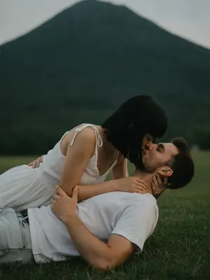 На фотографии в крупном плане запечатлены двое на фоне зеленой горы. Лежа на траве, они обнимаются и целуются, создавая интимное и непринужденное настроение. Женщина одета в воздушное платье с рюшами, а мужчина - в простую футболку и джинсы. Их руки ласково обняли друг друга, выражая нежность и близость.

Фотография пропитана мягким светом и теплой атмосферой летнего вечера. Тени идеально дополняют образы, создавая кинематографический эффект. Фон нежно размыт, а кожа выглядит реалистично, словно настоящая. Вся сцена словно застыла в случайно пойманном моменте, словно кадр из фильма.