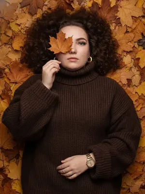 Фотореалистичное изображение, сделанное на iPhone. На плотном ковре из сухих кленовых листьев, окрашенных в золотистые, охристые, медные и коричневые тона, лежит женщина. Камера расположена чуть сбоку от ее тела, под углом, что позволяет увидеть ее голову в верхней части кадра, а корпус уходит вниз. Снимок близкий - женщина занимает почти все пространство, видна от макушки до середины груди, по краям остаются лишь немного листьев. Она расслаблена, голова слегка наклонена, лицо не строго анфас: подбородок повернут вбок, а взгляд направлен в объектив. Одна рука поднята, согнута в локте, и ее тонкие пальцы держат коричневый кленовый лист, который закрывает один глаз, в то время как второй глаз открытый и выразительный. На ней объемный темно-коричневый свитер оверсайз с крупной вязкой, свободно сидящий на плечах и корпусе, с высоким широким воротом и длинными рукавами. На поднятой руке, поверх свитера, находятся золотые часы с массивным металлическим браслетом, без дополнительных украшений. В ушах крупные эффектные круглые золотые серьги. Макияж включает длинные объемные ресницы с 2D-эффектом, а кожа имеет естественную текстуру с видимыми порами, без ретуши. Фон полностью состоит из осенних листьев, без лишних объектов, с немного пониженной экспозицией, натуральной цветопередачей и высокой детализацией, не размыт.