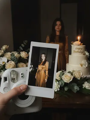 Transform the original image into a luxurious birthday party setting, maintaining the person's unique features and identity. The scene should exude an intimate, candid vibe, reminiscent of a spontaneous snapshot taken with an iPhone in dim lighting. The focus should be on a white instant camera capturing a freshly printed polaroid photo, held by a natural-looking hand with polished nails if suitable.

The polaroid should display the person elegantly posing at their birthday celebration, exuding confidence and refinement. Their outfit should be transformed into a polished, gender-appropriate birthday look that complements the original reference photo. In the background, a subtly blurred image of the person in the same outfit should be visible, creating a harmonious composition.

Surrounding the scene, include tasteful birthday decorations such as elegant flowers that match the subject's outfit and the overall color scheme. A birthday cake with a single lit candle, adorned with neat "Happy Birthday" icing, should be placed to the side. The lighting should be a mix of direct flash highlights, warm candle glow, and soft party lights, enhancing the atmosphere of the luxury birthday setting.

Maintain a realistic aesthetic with attention to detail, ensuring no changes to the person's facial structure, identity consistency between the polaroid and background, or deviations from the elegant and cohesive color palette of the original reference image.