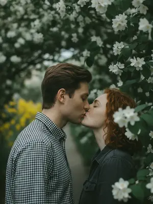 A woman and a man are locked in a tender kiss, their profiles touching, eyes shut in a moment of intimacy. The camera captures them up close, from shoulders to heads, with the focus at eye level, surrounded by blooming boughs. The foreground is a blur of branches and flowers, framing their faces at the center of the shot.

Their skin looks flawlessly smooth and matte, basking in the gentle light. The man sports a black and white checkered cotton shirt, while the woman is clad in a dark gray jacket.

The setting is beneath a blossoming tree, its dense branches adorned with large white flowers and yellow stamens, creating a natural archway around the couple. In the background, yellow blooms add a touch of color to the scene.

The lighting is soft and even, with no distinct source, casting a muted, cool tone over the frame. Shadows are almost non-existent, and the green and white hues dominate. The skin glows subtly in the subdued highlights, complemented by the pronounced film grain.

Shot with an 85mm lens, the focus is razor-sharp on their lips and the meeting point of their faces, blurring out the man's shoulder and nearby branches in the foreground. The background foliage melts into bokeh, enhancing the dreamy, romantic atmosphere of the scene.

The overall aesthetic is reminiscent of a Candid Film portrait, capturing the essence of a soft, spring day with a botanical backdrop. The color palette is dominated by white blossoms, deep greens, and dark charcoals, all bathed in a cool, diffused light. The film grain adds a touch of nostalgia, while the shallow depth of field creates a sense of intimacy and focus on the couple's connection.