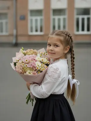Ребенок стоит на улице, повернув голову через плечо к камере, взгляд прямо в объектив, улыбается милой улыбкой. Ладонью касается края шапки, другой рукой прижимает к груди огромный букет цветов. Фото сделано снизу вверх, сосредотачиваясь на лице. Букет громадный, обернутый в мягкую розовую бумагу. В нем нежные гортензии, эустомы и ромашковые хризантемы различных оттенков. На заднем плане виднеется школьное здание. Освещение дневное, изображение зернистое. Фото высокого разрешения, живое изображение, как будто в 8К.