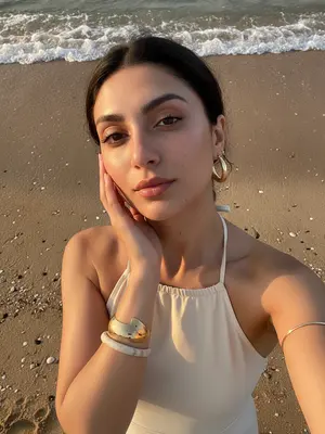 A woman stands gracefully on the sandy beach by the water, capturing a lifestyle beauty moment with her smartphone. The camera looks down on her, giving a high perspective typical of a handheld selfie. She tilts her head slightly upwards, gazing at the camera, while her hand delicately brushes the side of her face, fingers tracing the curve of her cheek and jawline. Her shoulders are relaxed, creating an elegant pose in the frame that captures her from the chest up.

Dressed in a cream halter neck swimsuit or summer top, her open shoulders and exposed collarbones exude a sense of summer chic. Adorning her look are large gold hoop earrings, a thick sculptural gold bracelet, a light marble-style bangle, and a delicate metallic arm bracelet above her elbow. Her makeup is soft and glamorous, perfect for a beach setting: her skin glows with a warm sun-kissed radiance, peach blush accentuates her cheekbones, clean sculpted brows frame her eyes, subtle winged eyeliner enhances her gaze, long lashes flutter, and her lips shimmer with satin finish.

In the background, the wet beach sand offers natural textures, with a thin line of sea foam marking the approach of a small wave behind her. Small shells are scattered on the sand, adding to the coastal atmosphere. The lighting captures the magic of a sunset or golden hour, casting warm highlights on her skin and creating soft shadows that play around her facial features. The shallow depth of field adds to the candid summer vacation mood, blending a realistic DSLR smartphone hybrid aesthetic seamlessly.