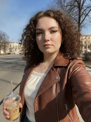 На ярком солнечном снимке запечатлена женщина, словно вдохновленная весенними прогулками. Она стоит на фоне ясного голубого неба и деревьев, лучи солнца играют на ее коже. Ее макияж аккуратен, с фокусом на сверкающих губах и четко выделенных бровях. Ресницы выглядят пышными, словно у лисы, а скулы нежно подчеркнуты контурингом.

Женщина одета в стильную кожаную косуху цвета шоколада, которая придает ей неповторимый шарм. Под курткой просматривается лонгслив из вискозы в молочных тонах с открытыми плечами. На ушах блеснут серьги-гвоздики, а на шее сверкает тонкая цепочка с небольшим бриллиантом.

Яркий солнечный свет создает игру контрастов и подчеркивает текстуру одежды и аксессуаров. Фон состоит из ясного голубого неба, обнаженных ветвей деревьев и асфальтного тротуара. Вдали виднеются изящные архитектурные сооружения, а женщина, словно модель, грациозно прогуливается по улице города.

Фотография сделана снизу вверх, на вытянутой руке, захватывая всю ее привлекательность и уверенность. Общее впечатление от снимка - позитивное, наполненное прогулочным настроением.