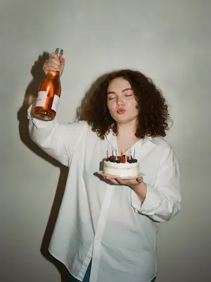 Изображение в стиле Party Girl / Birthday Aesthetic, сделанное в ультрареалистичной манере, как будто случайный снимок на старую цифровую камеру. Девушка в свободной белой рубашке оверсайз, застегнутой на пуговицы. В ее одной руке бутылка розового шампанского, которую она держит высоко, а другая рука держит небольшой бенто-торт с яркими свечами и свежими ягодами.