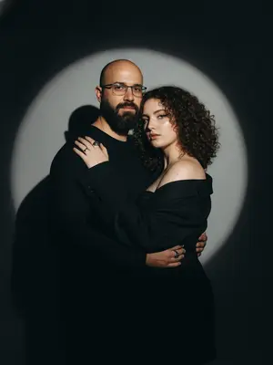 В студийном свете создан фотореалистичный портрет пары, олицетворяющий драматичную Y2K fashion-эстетику. Мужчина стоит на переднем плане, его корпус легко повернут в сторону, выставляя одно плечо ближе к камере. Поза его наполнена спокойствием и уверенностью, в то время как женщина стоит немного ниже, плавно обнимая его за шею и плечо. Ее лицо, выражающее дерзость и интимность, видно из-за его плеча, создавая эффект editorial стиля.

Женщина выглядит выразительно и гламурно, с cat-eye макияжем, четко очерченными губами и хайлайтером на коже. Она одета в черный корсет и оверсайз-пиджак с открытым плечом, в то время как мужчина носит черный лонгслив. Фон студии окрашен в темные оттенки, практически черный, с мощным прожектором, создающим большой круг света за спинами персонажей.

Свет от прожектора мягко освещает фон, растворяясь по краям и погружаясь в темноту, а персонажи частично перекрывают его, проецируя жесткие тени на стену. Освещение фронтальное, с высоким контрастом, реалистичными бликами и глубокими тенями на коже, а также легким пленочным зерном. Камера уровня глаз, с портретным объективом и плотным кадрированием, захватывает каждую деталь в 8K RAW качестве.

На снимке отсутствуют лишние элементы, текст и логотипы, подчеркивая исключительно модные образы и драматичную атмосферу.