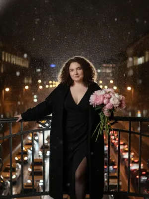 Портрет женщины в кинематографическом стиле. Снег и дождь смешиваются, падая на нее, стоящую на мосту в ночное время. Она одета в черное платье с открытыми плечами и глубоким вырезом, с высоким разрезом на ноге. Черные колготки из капрона и длинное пальто-накидку, расстегнутое на пуговицах. Макияж безупречен, улыбка нежная, взгляд устремлен прямо в объектив. Рядом с круглым железным поручнем моста она стоит, одна рука вытянута в сторону, в другой держит букет цветов. Вдалеке за ее спиной город ночной и дождливый, огни в окнах домов мерцают, под мостом машины стоят в пробке, фары сверкают. Фотография с использованием вспышки, с эффектом зернистости, создает атмосферу загадочности и романтики.