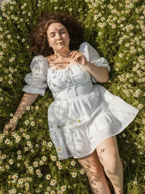 Captured with the latest iPhone 16 Pro Max, the device discreetly absent from view. A strikingly authentic photo, devoid of artificial enhancements, showcasing the woman's natural pores and skin texture. She reclines gracefully amidst a lush field of chamomile, the camera directly above, capturing her from head to knees in a diagonal composition - head angled towards the upper left, legs extending to the lower right.

Enveloped by a sea of white chamomile blooms, her eyes peacefully shut, a soft, serene half-smile gracing her lips. One hand delicately lifts a chamomile stem to her face, the flower tenderly brushing against her lips. A tranquil, almost poetic ambiance fills the scene, accentuated by her hair splayed in a halo around her, mingling with the grass and blossoms, a few stray strands caressing her cheeks and forehead.

She wears an ultra-short, romantic peasant-style dress in pristine white, featuring billowy off-shoulder sleeves, a square neckline adorned with front ties, and a voluminous, layered skirt crafted from textured broderie anglaise cotton. A gentle breeze lifts the skirt slightly, revealing her bare shoulders, collarbones, and legs. A single chamomile blossom rests delicately on the fabric.

The bright, sunlit day casts intricate lace-like shadows across her attire, face, arms, and legs, the entire frame awash with the vibrant hues of green stems, wild grass, and the pure whites of the chamomile flowers. The scene exudes a countryside editorial aesthetic reminiscent of a Pinterest board, with natural iPhone colors enhancing the rich greens, clean whites, and warm daylight, subtly heightened by a touch of contrast.