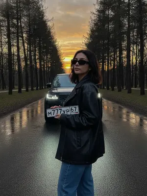 На заднем плане раскинулась пустая дорога в парке с атмосферным освещением закатного неба, его золотистые оттенки отражаются на мокром асфальте. Лазерные фары автомобиля без номеров добавляют кинематографическую атмосферу, создавая luxury automotive mood с низкого угла обзора.

Модель стоит рядом с машиной, держа перед собой номерной знак. Она надевает oversized черную кожаную ретро-куртку и джинсы, а на лице - прямоугольные черные солнцезащитные очки в металлической оправе. Уверенная и расслабленная поза модели добавляет нотку стиля к кадру.

Фотография зафиксирована на уровне пояса, сосредотачивая внимание на модели и автомобиле. Задний план занимают отражения деревьев и неба, создавая гиперреалистичный эффект закатного света. Съемка выполнена с использованием камеры Sony A7R IV и объектива 85мм, обеспечивая ультрачеткое изображение с неглубокой глубиной резкости.

Избегая негативных аспектов, таких как размытость, искаженный автомобиль, фальшивые отражения и плохие руки, фотограф стремился к созданию идеальной кинематографической сцены. На снимке отсутствуют лишние элементы, перегруженный текст и насыщенные цвета, что подчеркивает реализм и эстетику момента.