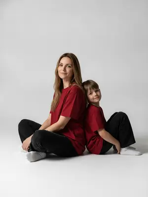 Изображение с высоким разрешением, точностью в 8K. На фото запечатлены женщина и ребенок, одетые в красные свободные футболки и черные широкие джинсы, а также в белые носки. Женщина сидит на полу, согнув колени, с улыбкой на лице, направляя свой взгляд прямо в объектив. За ее спиной, в той же позе, сидит ребенок, слегка улыбаясь, опираясь спиной о спину женщины. Освещение студийное, с глубокими тенями, создающее драматический эффект. Фоном служит чисто белая плоскость, подчеркивающая элегантность и силу композиции. Изображение выполнено в темном стиле модного журнала, с ультра-реалистичным подходом и высокой степенью детализации. Портрет выдержан в художественном ключе, придавая ему особую выразительность.
