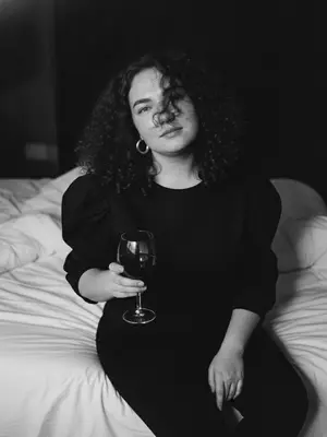 Capture a black-and-white portrait in the Y2K style, taken at night with a direct flash. The subject, a woman, sits gracefully on a tousled white bed, viewed from a slightly raised perspective. She is adorned in a chic black top featuring billowy sleeves and striking circular earrings. One hand delicately cradles a sophisticated glass of deep red wine. Her body subtly sways, imparting a natural blur to her face and shoulders, as if frozen mid-movement. Her gaze is tender, meeting the camera with a soft, intimate smile.

Flowing locks cascade in loose waves, with stray strands gently brushing her face, tousled by her motion. The lighting is stark, casting sharp shadows against the night backdrop, creating a high-contrast ambiance with intense highlights and deep shadows, accentuated by visible film grain. Her skin appears authentic and textured, untouched by digital smoothing, enhanced by the flash's glare.

The overall atmosphere exudes a high-fashion editorial vibe, blending Parisian elegance with a sense of old-world allure. The scene evokes a melancholic nighttime sentiment, capturing a spontaneous and cinematic essence. Shot in a raw 35mm film style, the image boasts pronounced motion blur, adding a touch of lived-in realism to the captivating composition.