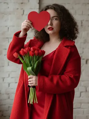 На высокоразрешенном фото моды женщина сжимает букет красных тюльпанов, закрывая один глаз крупным красным сердцем. Она надевает элегантное красное платье на тонких бретельках и оверсайз красное пальто с текстурой. Лицо модели освещено мягким светом слева, создавая нежные тени. Взгляд игривый, губы ярко-красные, лицо выдержано в точных чертах. Фото в стиле журнального бьюти, с романтическим настроением и теплой красно-зеленой палитрой. Фон - белая кирпичная стена, добавляющая шарму. Кадр в трех четвертях с небольшим наклоном вверх, с четким фокусом на лице и небольшой глубиной резкости. Изображение супердетализировано, в формате 8K, идеально подчеркивающее красоту и стиль модели.
