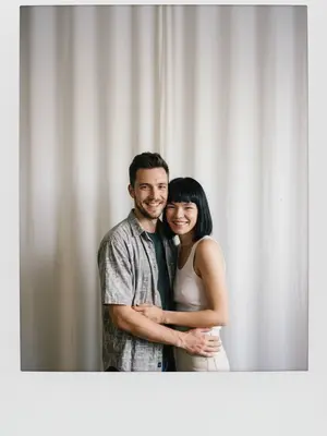 Сделайте снимок с помощью фотоаппарата Polaroid. Фотография должна выглядеть как обычное фото, без четкого объекта или реквизита. На фото должен быть постоянный источник света, такой как вспышка из темной комнаты, равномерно распределенная по всей фотографии. Не меняйте лица. Замените фон за спинами двух человек на белую занавеску. Люди на фотографии обнимаются.