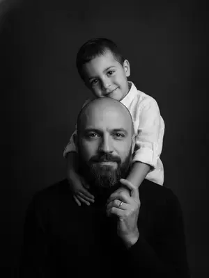Изображение показывает родителя и ребенка, которые участвуют в семейной фотосессии. Фото выполнено в студии и имеет вертикальную композицию, с крупным планом лиц. Родитель, находящийся внизу кадра, направляет свой взгляд прямо в объектив, лицо полностью обращено к нам. Ребенок, находящийся сверху, повернул голову в сторону, его щека лежит на плече родителя, и он смотрит в камеру с нежной улыбкой. Рука ребенка обнимает шею родителя, видна в кадре, а родитель, в свою очередь, держит эту руку своей ладонью.

Одежда ребенка состоит из белой рубашки из льна, в то время как родитель одет в черную водолазку. Фон студии выполнен в темно-серых почти черных тонах, что создает контраст с яркими лицами на переднем плане. Освещение мягкое и боковое, оно придает изображению объем и глубину.

Фотография выполнена в черно-белой гамме с выразительными контрастами. Атмосфера, царящая на снимке, пропитана нежностью, интимностью и спокойствием.
