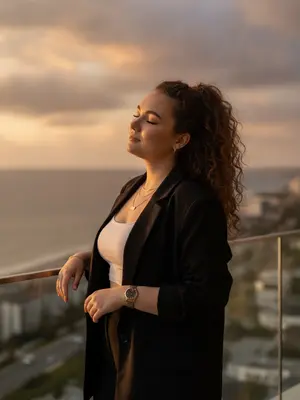 A woman stands gracefully on a balcony overlooking the ocean, bathed in the warm glow of the setting sun. Her body is slightly angled, one arm casually resting on the glass railing as she basks in the sunlight with her eyes closed, exuding an air of effortless elegance.

She is dressed in an oversized black blazer paired with a fitted white crop top and elegant black trousers, adorned with minimal yet luxurious jewelry and a sleek watch that adds a touch of old-world sophistication to her ensemble.

Her makeup is a soft glam look, enhancing her bronzed skin with a natural contour, defined brows, and long, fluttering lashes. The warm sunlight accentuates the realistic texture of her skin, casting soft highlights that add to her radiant glow.

The lighting captures the golden hour ambiance of the sunset, creating a cinematic glow that bathes her in a warm, ethereal light. The soft shadows and highlights play delicately on her hair and skin, enveloping her in an aura of luxury and tranquility.

In the background, the ocean stretches out beneath a soft, cloudy sky, with the city's coastline and roads below blurred into a dreamy backdrop that adds depth to the scene. The overall atmosphere is one of relaxation and sophistication, capturing a moment of pure serenity and beauty.

The image is captured with an iPhone, giving it a realistic and candid feel with a 50mm lens look that focuses on the woman while softly blurring the background. The emphasis is on creating an ultra-realistic portrait that exudes a luxury influencer vibe, showcasing natural body proportions and skin texture with a film-like quality that avoids overprocessed artificial enhancements.