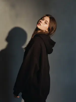 На съемке haute couture запечатлена стильная женщина, стоящая спиной к наблюдателю. Ее голова наклонена назад, взгляд устремлен прямо в объектив. Легкая улыбка играет на ее губах. Фотография сделана на 35-мм пленку с высоким контрастом, создавая жесткое драматическое освещение. Резкая тень отчетливо отражается на простом сером фоне, придающем образу эстетику 90-х. Теплая коричневая цветовая гамма добавляет кинематографичности и стиля, характерного для фэшн-журналов.

Изображение характеризуется максимальным фотореализмом, чистой редакционной фотографией с высоким контрастом и четкими контурами силуэта. Естественная анатомия, реалистичные руки без лишних пальцев, никаких дополнительных конечностей или дублированных частей тела. Лицо не искажено, не имеет деформаций. Нет запутанных нитей, сеток, текста или водяных знаков. Фотография выполнена в цвете. 