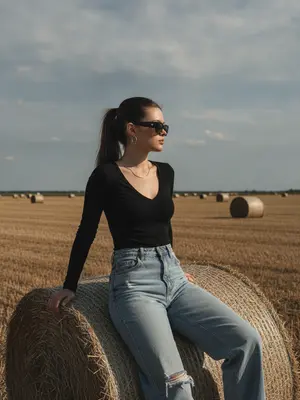 На фотографии в стиле high-fashion editorial, женщина сидит на большом тюке сена в сельской местности. Ее корпус слегка повернут, одна нога свободно опущена, а другая согнута, создавая расслабленную, но выразительную позу. Руки лежат естественно на колене и поверхности тюка. Взгляд направлен в сторону, профиль лица выражает спокойствие и уверенность. Женщина одета в черный топ с V-образным вырезом и объемные светлые джинсы с потертостями. На ней крупные солнцезащитные очки и минималистичные украшения. Фон состоит из открытого поля с несколькими тюками сена, создающими перспективу. Линия горизонта находится ниже центра кадра, подчеркивая небо. Освещение жесткое, натуральное, смягченное облаками, создающее выразительные тени и контраст. Стиль фотографии кинематографичен, с пленочным зерном и высокой детализацией. Редакционный fashion стиль акцентирует внимание на атмосфере и пространстве кадра. 