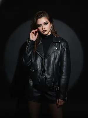 На стильной студийной сцене в эстетике "fashion editorial" запечатлена дерзкая фотография.
Женщина, стоящая в центре кадра, излучает уверенность. Одна из ее рук легко прикасается к лицу, задевая серьгу, вторая же расслаблена. Ее взгляд пронзителен и прямой, выражая холодную уверенность и самообладание.
Она одета в черную куртку-косуху оверсайз с металлической фурнитурой, которая перекидывается поверх черной водолазки. Кожаные шорты с высокой талией и ремнем, а также черные полупрозрачные капроновые колготки дополняют ее образ. Ткань куртки имеет матовый блеск и естественные складки на сгибах.
Фотосессия проходит в минималистичной студии с черным фоном, на котором ярко выделяется круглый световой пятно.
Освещение контрастное, напоминающее спотлайт, направленное спереди. Свет создает четкий круглый ореол за спиной модели и бросает глубокую, резкую тень на фоне. Светотеневые игры видны на лице и одежде.
Профессиональная камера с объективом 85mm обеспечивает идеальные портретные пропорции без искажений.
Модель находится в резком фокусе, в то время как фон за пределами светового пятна погружен в черную пустоту.
Макияж модели драматичен: "smoky eyes" в темных тонах, четко очерченные брови и матовая помада.
Дополнительные детали включают ярко-красный маникюр на длинных ногтях, крупные золотые серьги-кольца, металлический блеск молний на куртке и тонкое кольцо на пальце.
Атмосфера фотосессии наполнена роковым, сильным и графичным настроением, подчеркнуто модным. 
