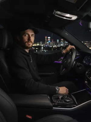 Мужчина с элегантным образом за рулем роскошного BMW, стоит на фоне ночного города. Его рубашка тщательно заправлена, подчеркивая уверенность и стиль. Серебряные часы на запястье и цепочка на шее добавляют шик. Ладонью касается руля, взгляд направлен прямо в объектив с уверенностью. Салон автомобиля пропитан атмосферой роскоши: кожаные сиденья и яркие фиолетовые светодиоды создают уют. Вдали сверкают огни городской ночи, придающие образу стиль, статус и мощь. Съемка осуществлена на объективе 50mm f/1.8 с использованием вспышки, игра света и тени придают изображению выразительность. 