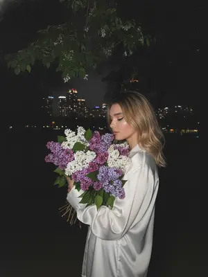 На темном фоне ночного города, тенистые деревья и майская листва создают задний план. Женщина стоит перед камерой, обе руки обнимают огромный букет сирени, белой и фиолетовой. Ее голова слегка наклонена к цветам, словно вдыхая их аромат. Макияж подчеркивает глаза черными стрелками и длинными ресницами, на губах блеск. Съемка происходит сбоку на уровне глаз, при слабом ночном освещении. Изображение высокой детализации, снятое на iPhone 17 Pro, передает живой момент с легким цифровым шумом и зернистостью, без использования вспышки. 