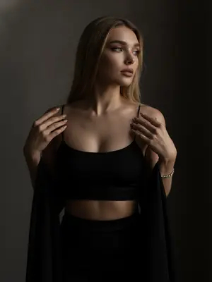 Фотография в стиле moody boudoir editorial, воплощающая атмосферу интимности, чувственности и приглушенного драматизма, представляет собой крупный план по пояс. Женщина стоит в полумраке, ладонями касается края черной ткани, изящно придерживая ее. Ее плечи слегка приподняты, а руки с длинным светлым маникюром сжимают тонкие бретели топа. Голова немного повернута в сторону, взгляд задумчиво устремлен вдаль, а выражение лица томное, загадочное и уверенное.

Вечерний макияж добавляет выразительности: гламурные стрелки, густые ресницы, создающие эффект «кошачьего взгляда», мягкий контуринг лица и матовая помада цвета пыльной розы. Женщина одета в облегающий черный кроп-топ на тонких бретелях-спагетти из матовой эластичной ткани, дополненный изящным серебряным браслетом на запястье.

Интерьер минималистичен, в темных тонах, с однотонным фоном, слегка текстурированным, погруженным в глубокую тень, создающую камерную обстановку. Низкоключевое освещение мягко падает сбоку, подчеркивая рельеф ключиц, линию плеч и текстуру кожи, создавая объем и драматичное настроение.

Используя объектив 85мм f/1.4, фотограф добивается идеальной фокусировки на глазах и лице модели, смазывая плечи и руки в мягком размытии. Гиперреалистичная текстура кожи, видимые поры, естественный блеск и отсутствие пластичности после ретуши создают естественный образ.

Общая эстетика фотографии - "dark feminine energy", создает интимную, теплую и дорогую атмосферу, наполненную мистикой и загадкой. 